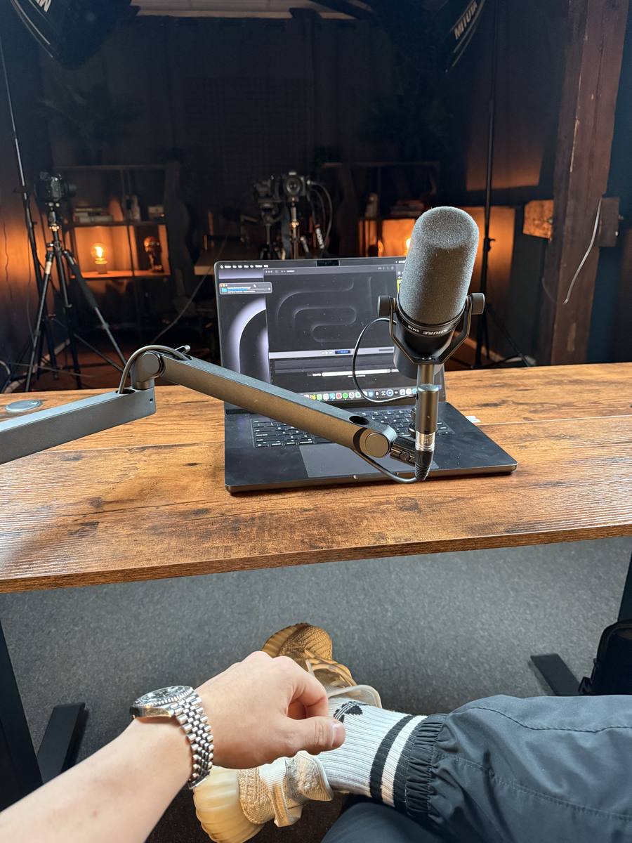 A studio microphone on a boom arm in front of an open laptop on a wooden desk, cameras and warm lamps behind